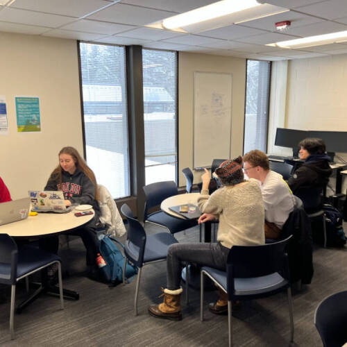 Students sitting in groups at tables in a bright classroom, working on laptops and engaging in discussion, with large windows, posters on the wall, and snacks on a side table